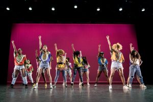 Group of female dancers standing with fists raised on stage against a purple backdrop.