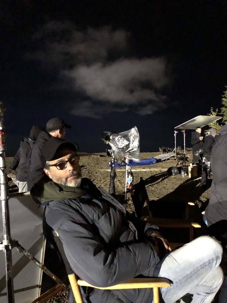 A man wearing a black puffer coat and ballcap is seated in a director's chair outside at night surrounded by camera filming equipment.
