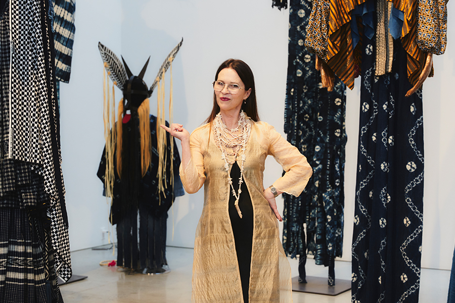 Laura Andeson Barbata standing in her studio, showing off some of her work, composed of costumes created from dark and textured fabrics and other materials.
