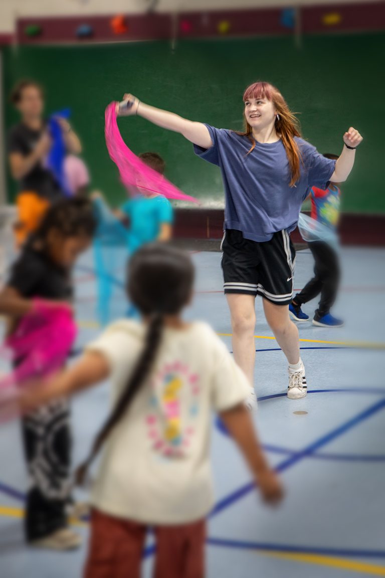 Dancer with pink hair and blue shirt surrounded by kids and waving a pink scarf.