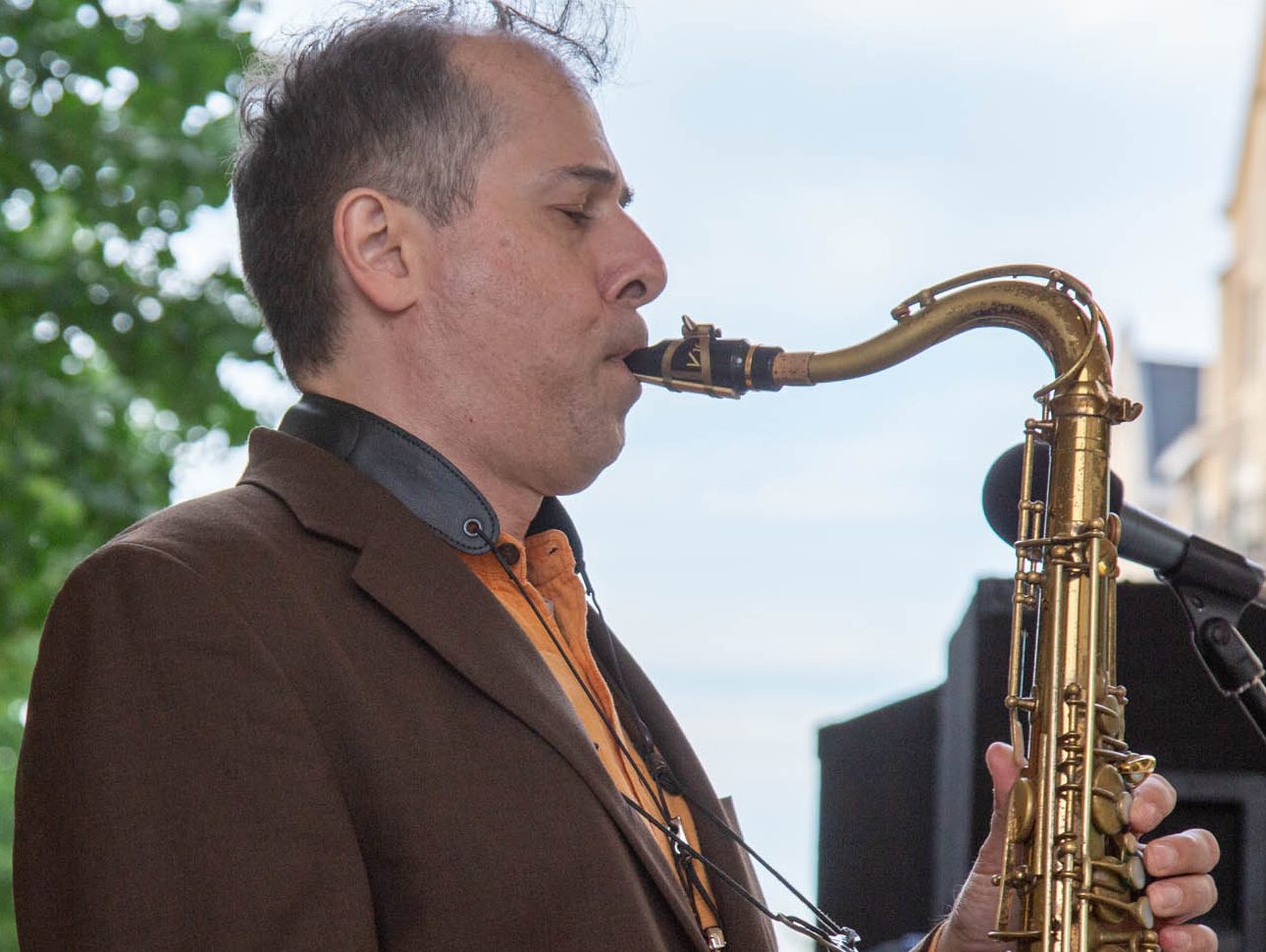A man in a brown suit jacket blows on a tenor saxophone.