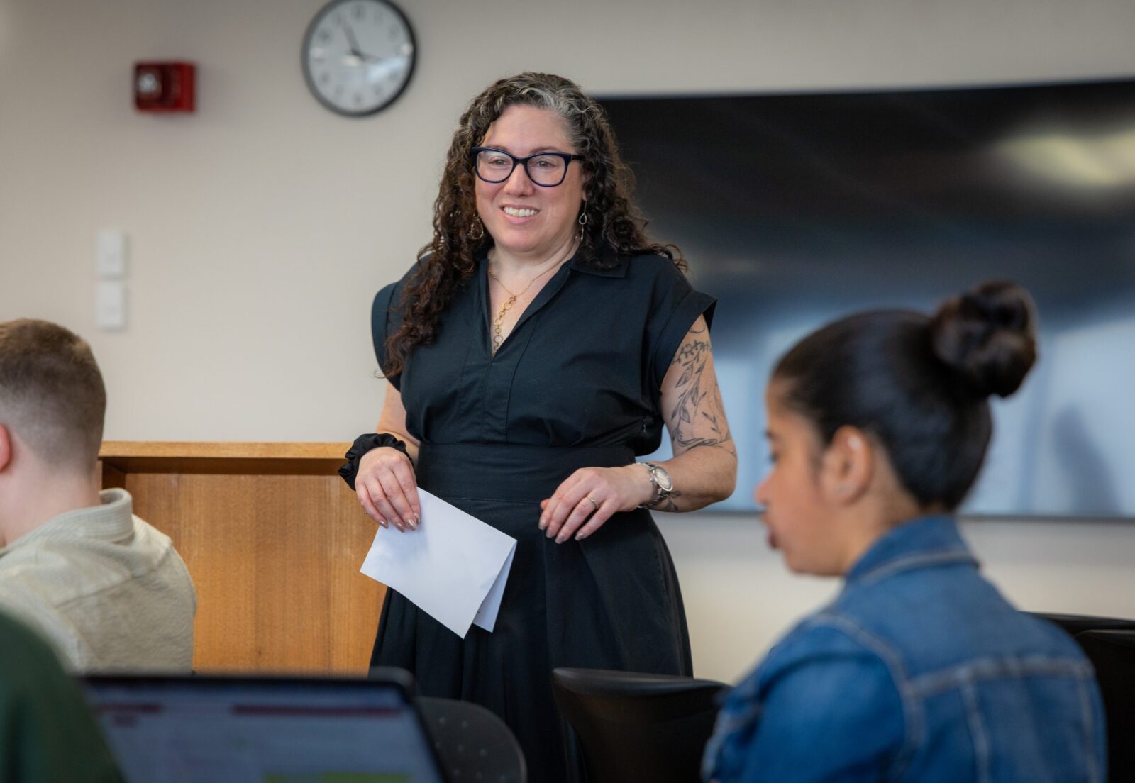A woman in black with long curly hair, glasses and a tattoo on her left arm stands in front of a class.
