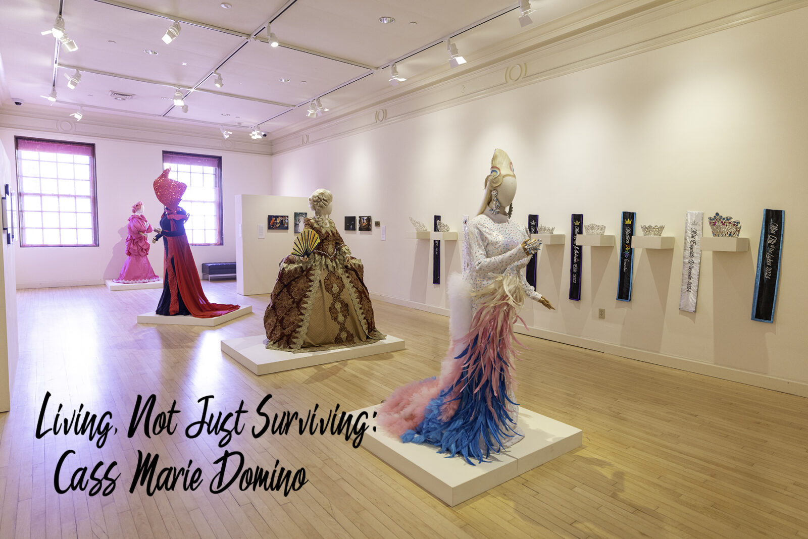 A wide shot of the gallery space with four mannequins in gowns backlit in pink. Text on the photo says "Living, Not Just Surviving: Cass Marie Domino"