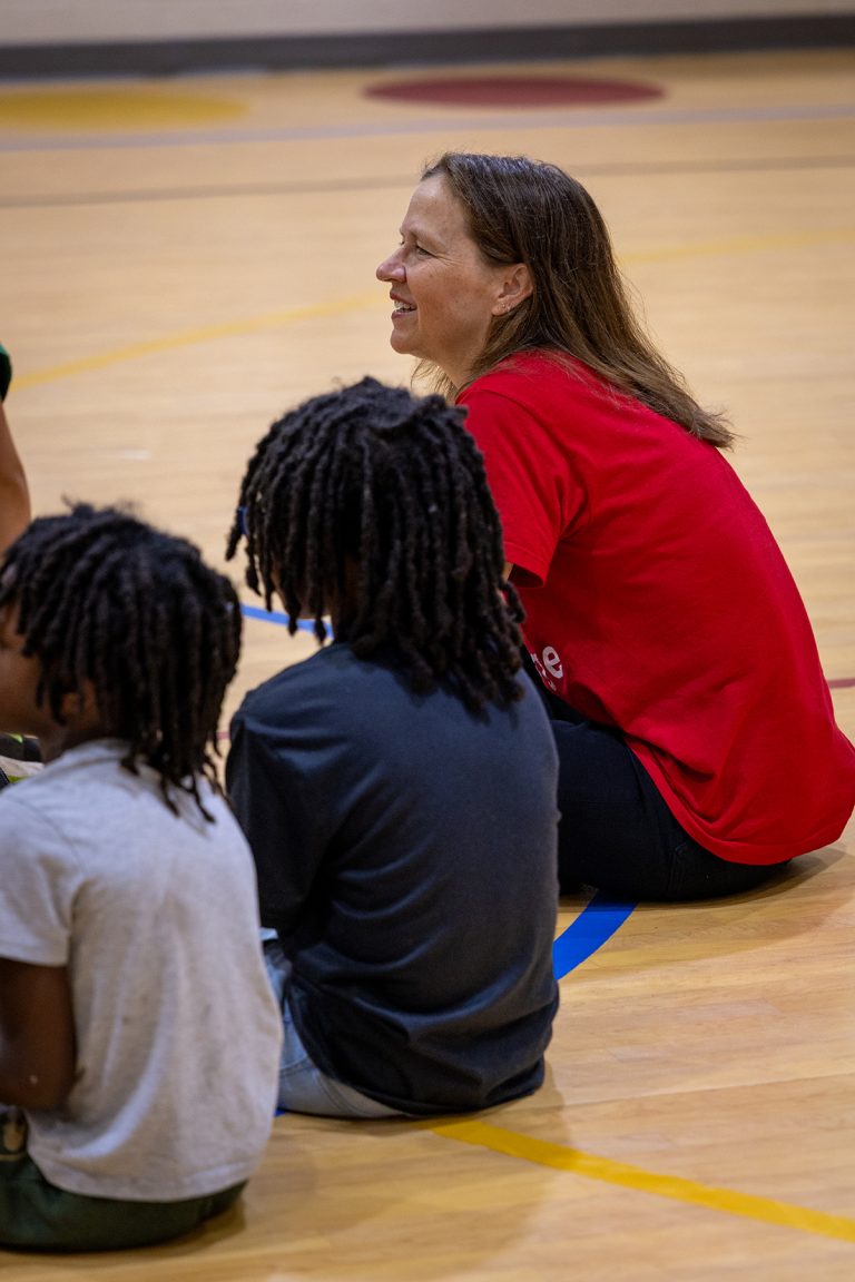 A woman in a red shirt sits in a circle with two children whose backs face the camera.