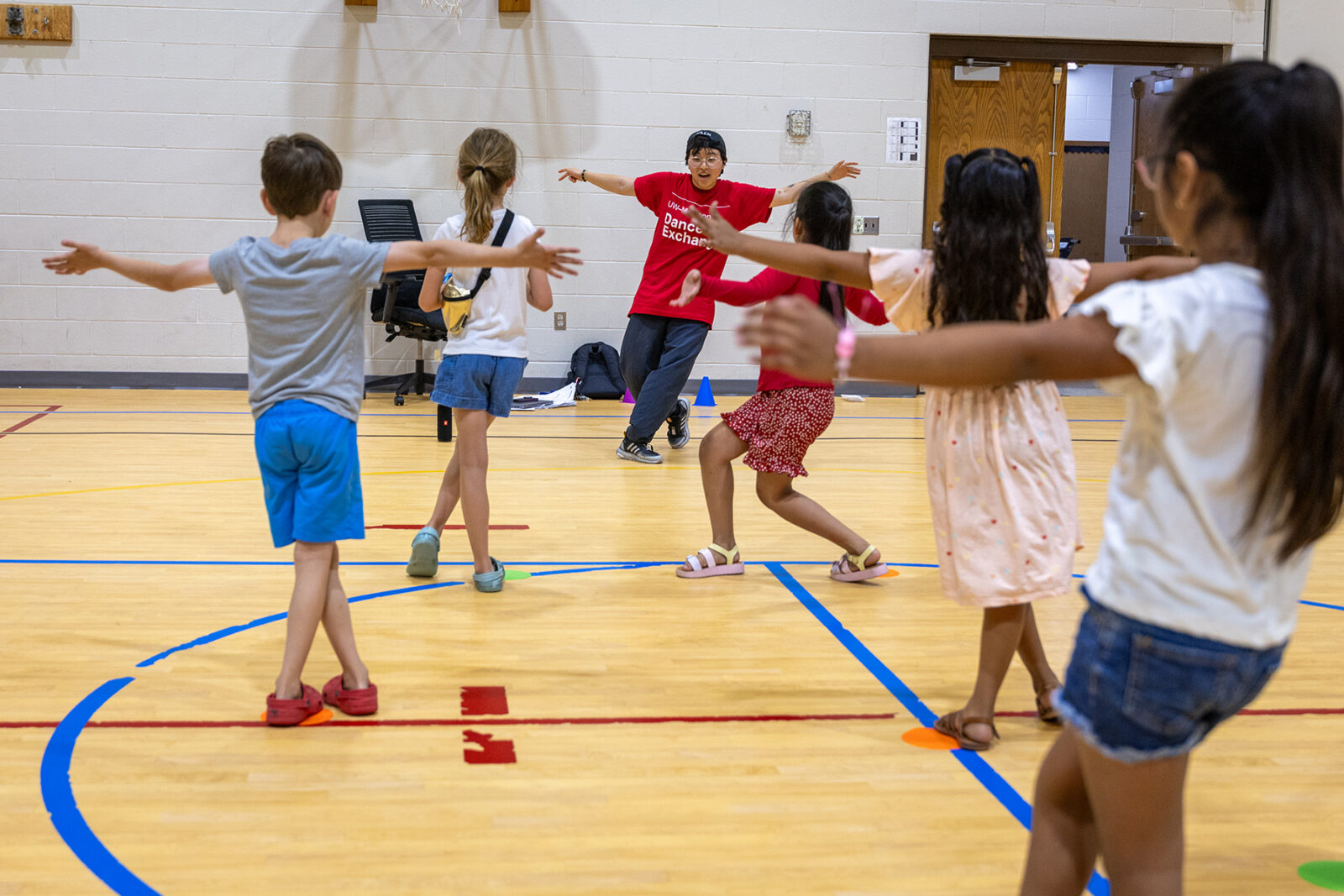A dance teacher with arms out wide instructs kids in school gymnasium.