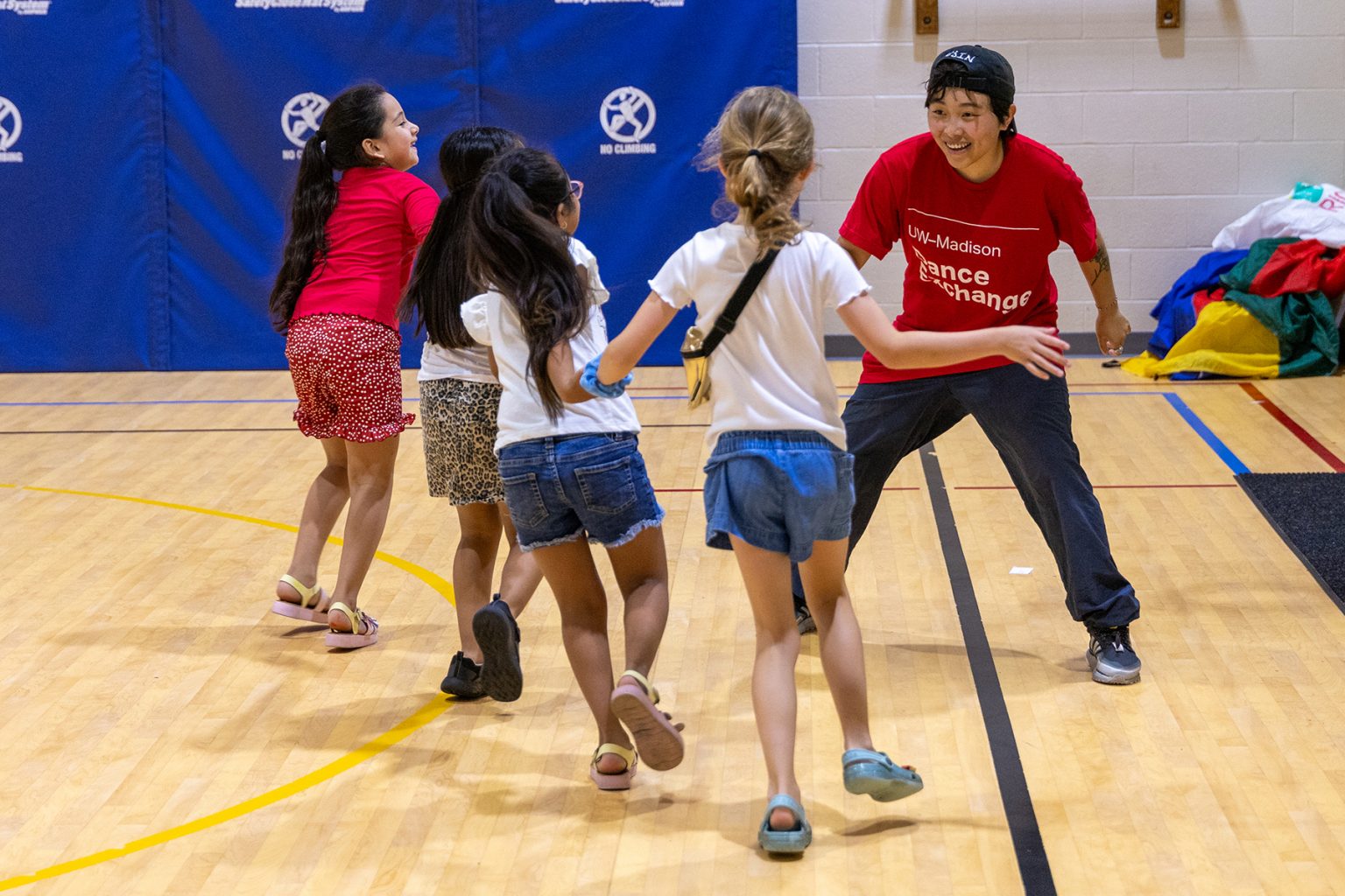 A teacher in a red shirt smiles as she watches four children run while holding hands.