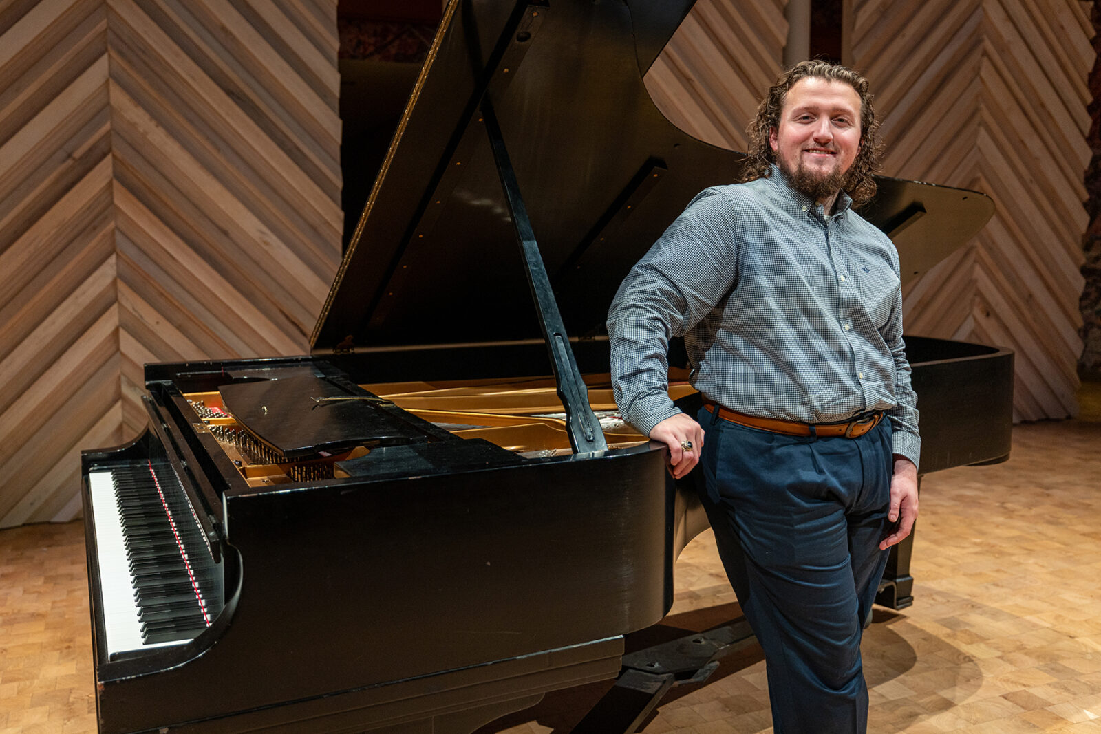 A man with curly hair and a beard leans against a concert Steinway piano on a stage.