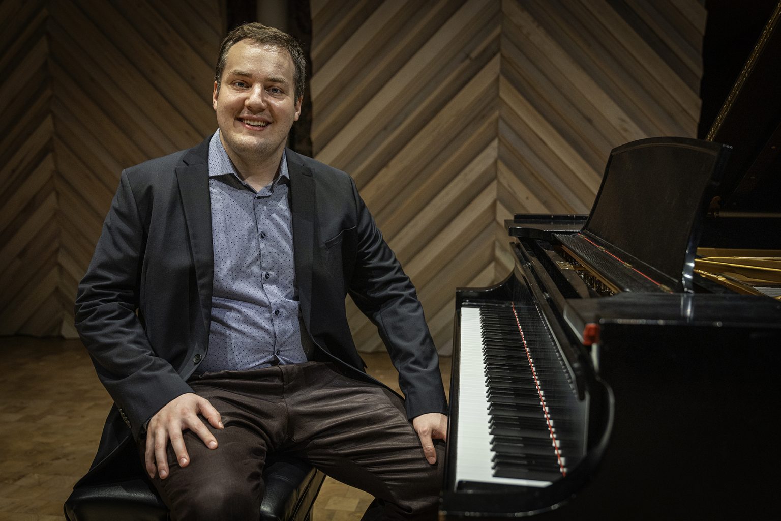 A man in a sport coat is seated smiling at a grand piano.