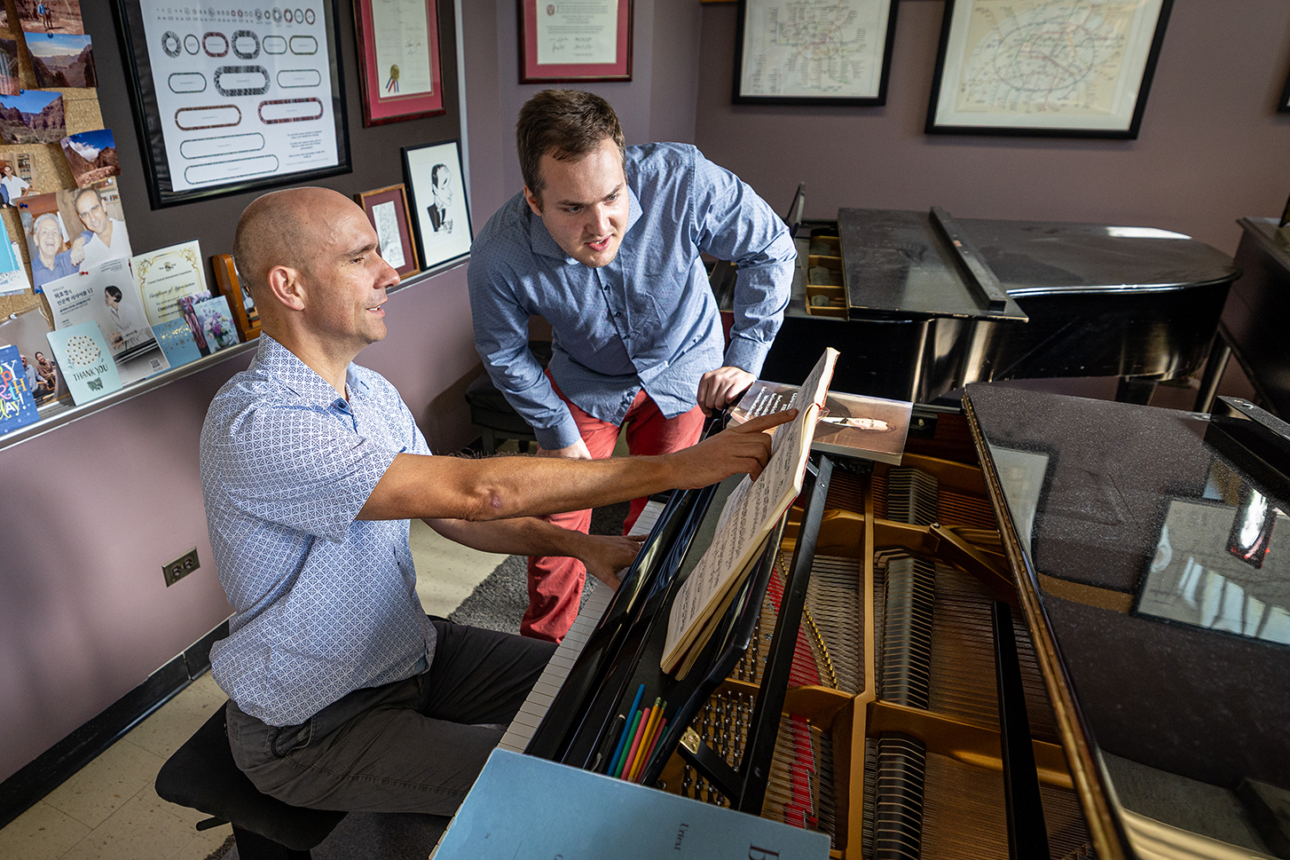 A man seated at the piano points to the sheet music as the student leans over to watch.