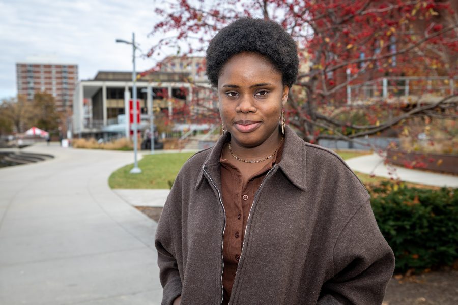 A woman dressed in a brown jacket smiles lightly at the camera outdoors with buildings behind her.