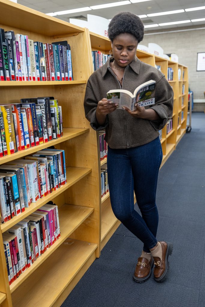 A woman stands reading a book leaning against the library bookshelves.