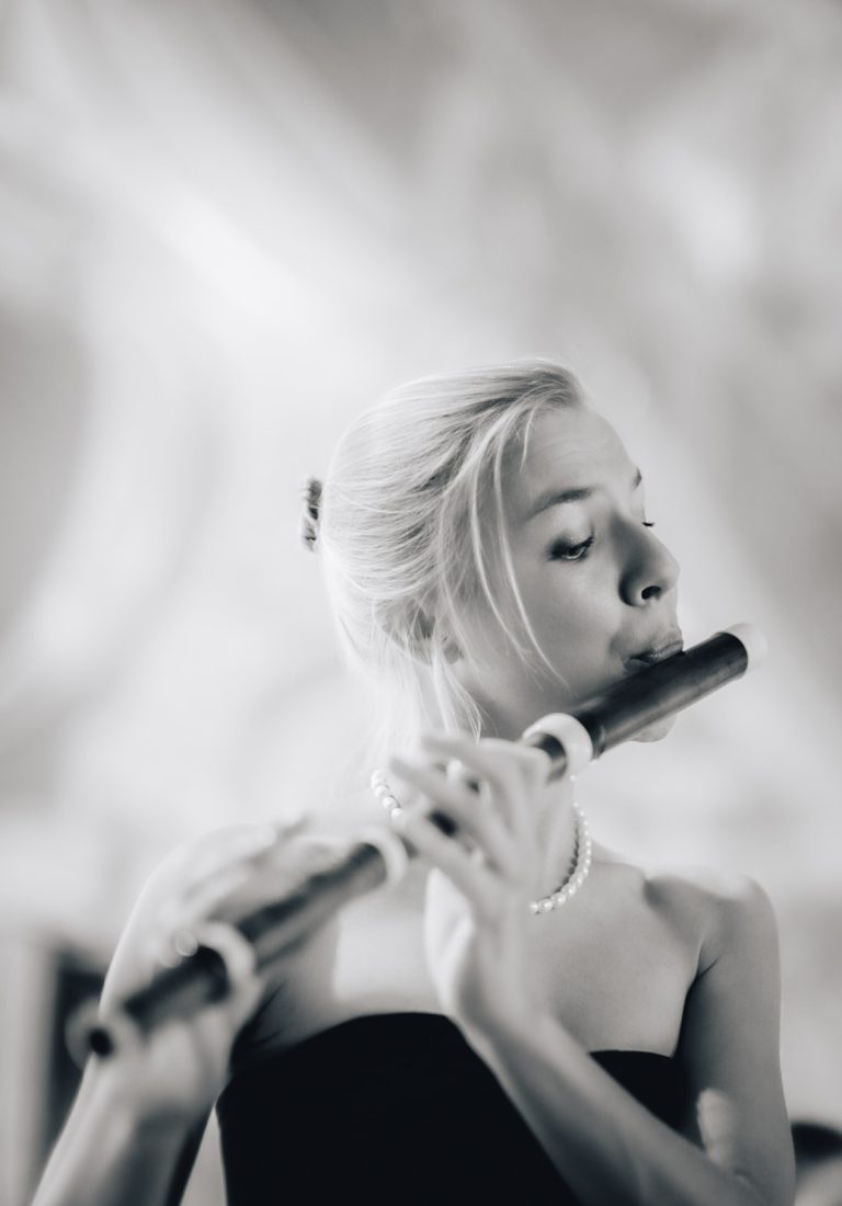 close-up black and white photo of Taya König-Tarasevich dressed in a black gown and white pearls playing a historical flute