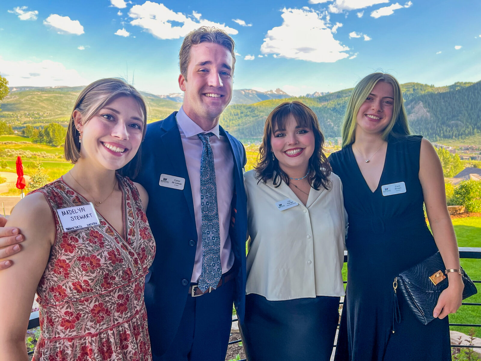Four people in dress attire stand in front of a mountainous background and blue skies.