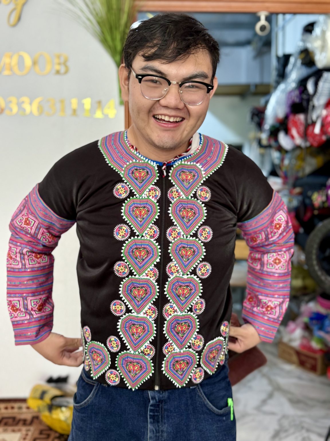 A male student wearing glasses and a traditional Hmong embroidered shirt smiles into the camera