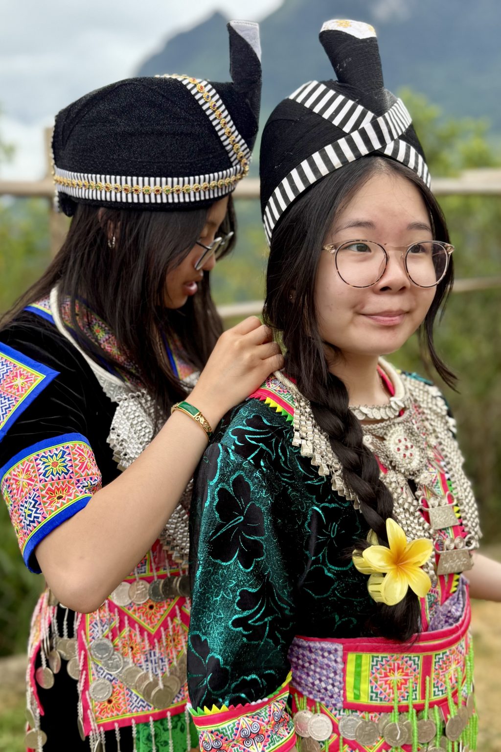 Two students stand in profile, both traditionally dressed in Hmong fashions, one standing behind the other pinning something on.
