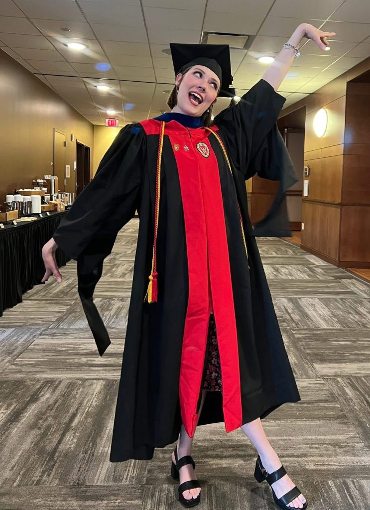 A woman dressed in a cap and gown poses in a hallway after a graduation ceremony.