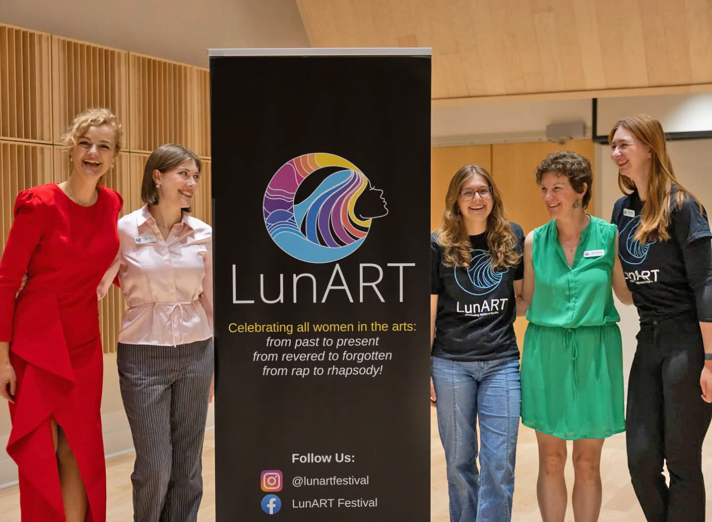 Five people in dress attire stand indoors around a big black sign with a colorful logo that says LunART.