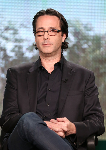 A man wearing a dark suit jacket an dark jeans sits in front of a window with trees visible behind him.