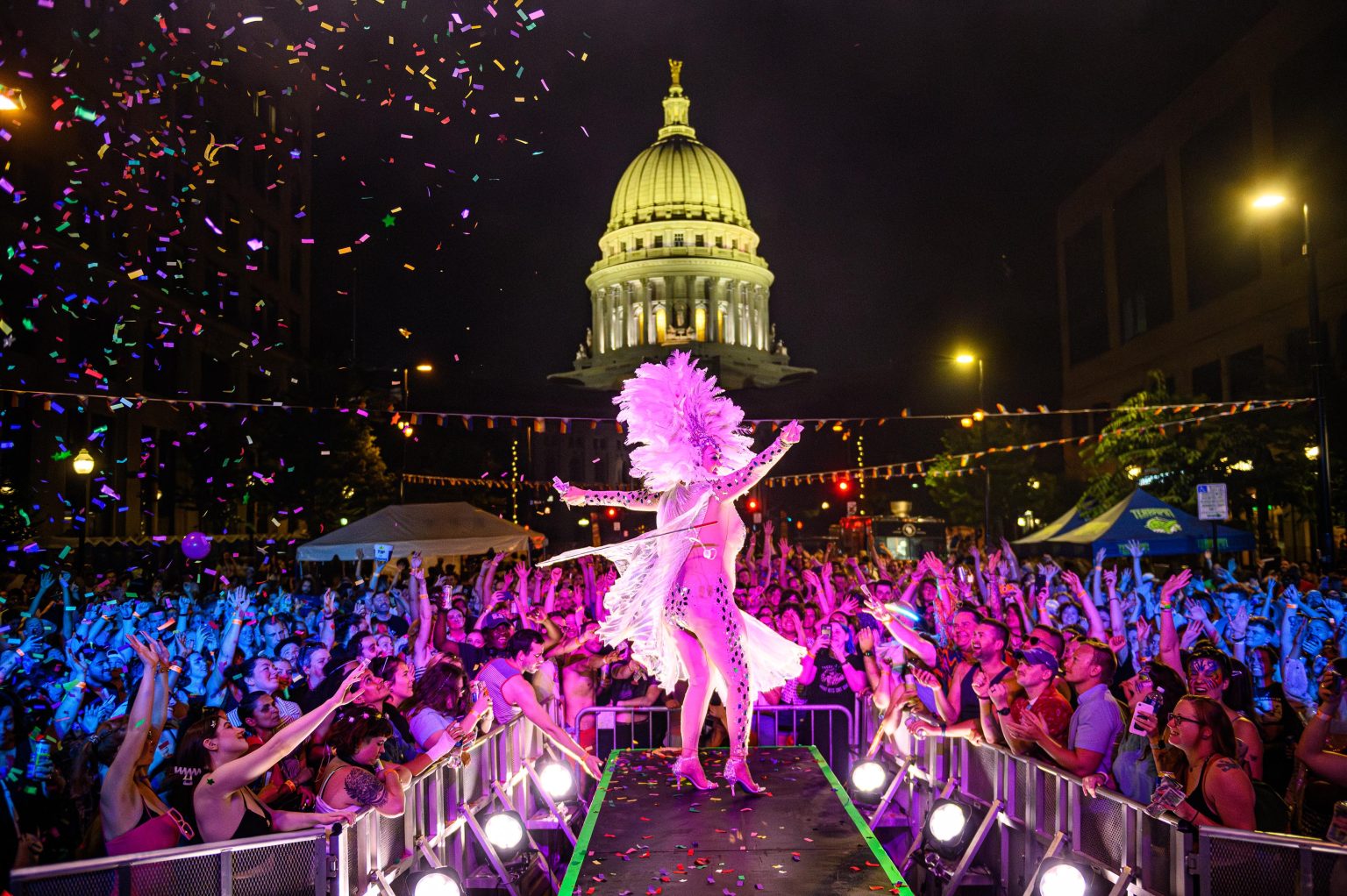 The drag queen stands on an outdoor runway in front of the capital dome at night in a crowd of people cheering.
