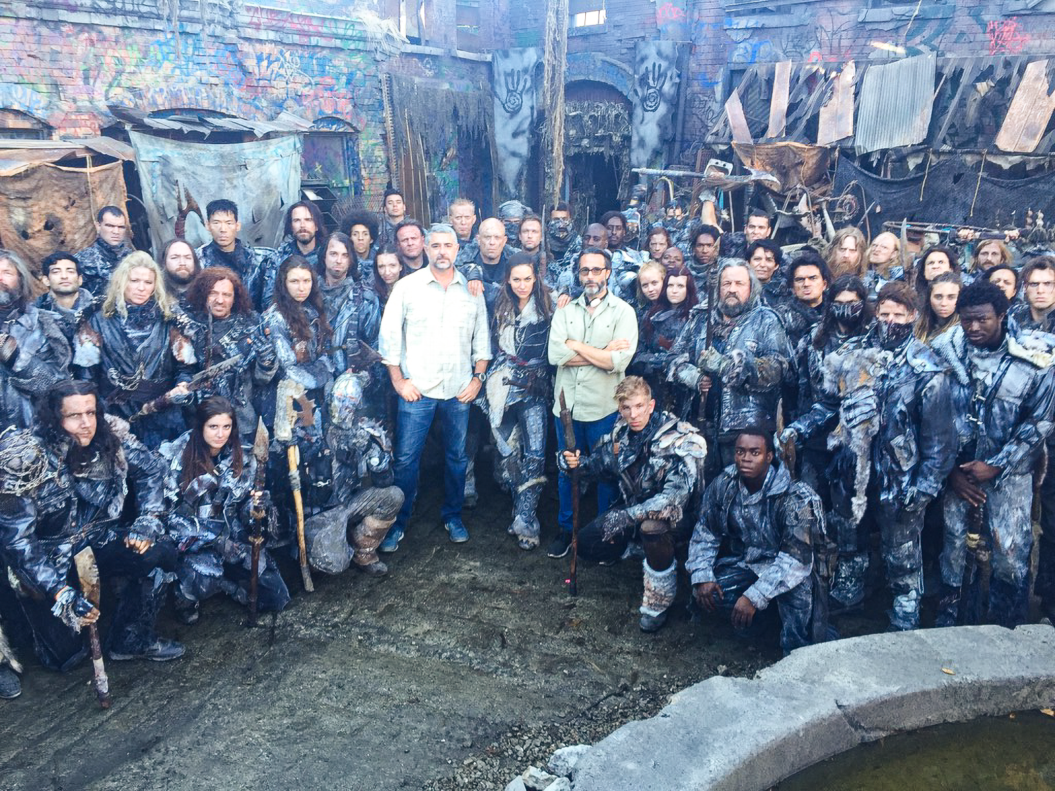 A large ensemble cast of The 100 in full costume is gathered on a futuristic-looking set with two director/producers standing in plainclothes at the center.