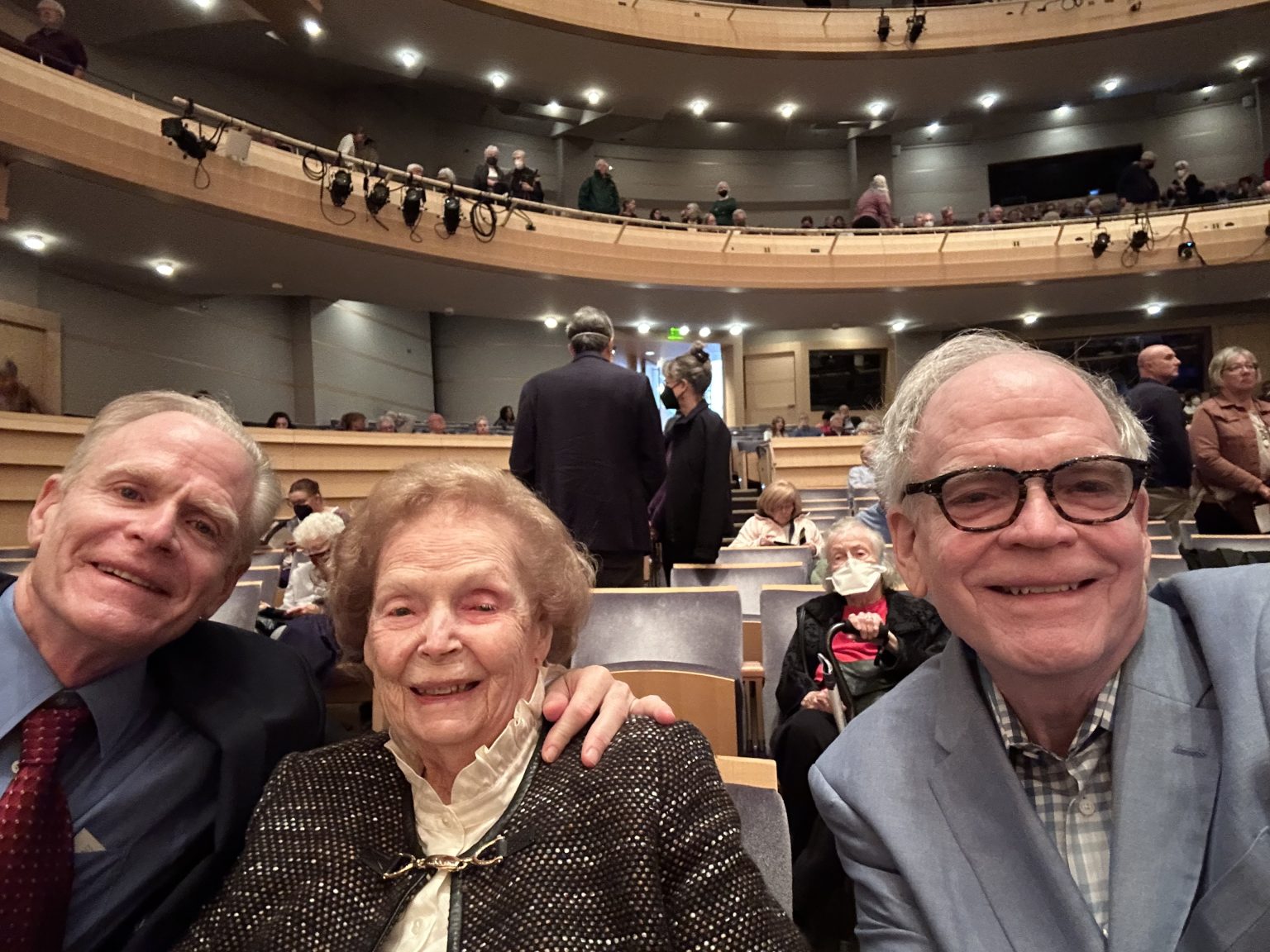 A woman seated in a concert hall with two grown sons, one on either side of her.