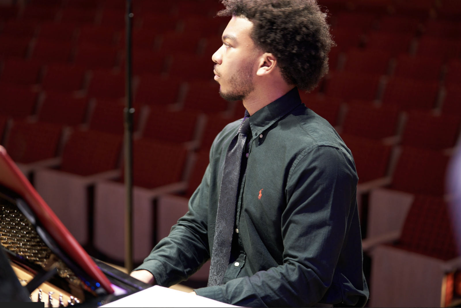 A student wearing a black collared shirt and black tie sits at a piano looking off to his right with empty concert seats behind him.