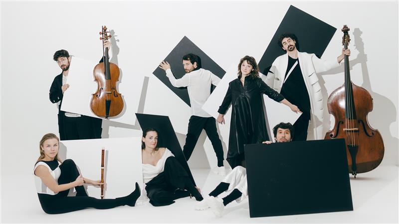 Seven members of Verità Baroque ensemble all dressed in black and white strike vivid poses with their instruments against a white background