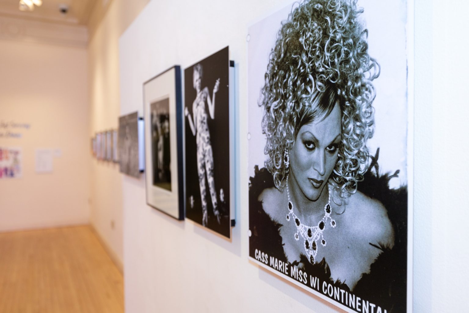 A close-up of a gallery wall where only one mounted black-and-white photo is in focus. The model has a big curly wig and the text says "Cass Marie Miss WI Continental"