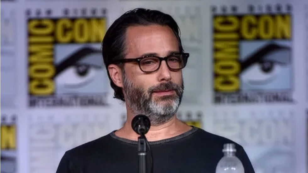 A man with dark hair, a salt and pepper beard, and dark glasses sits before a microphone with a comic con backdrop behind him