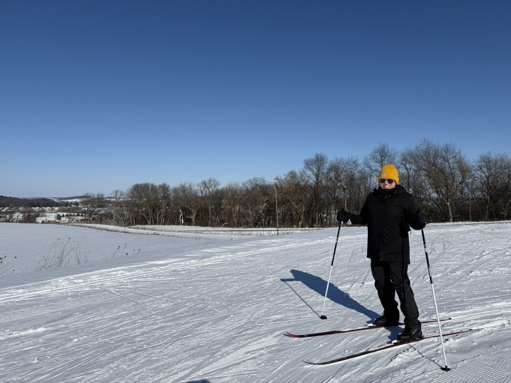 A bundled up woman in a yellow hat wearing skis and holding poles stands on a snowy spot.
