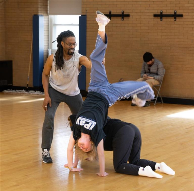image of dance instructor assisting one student tumble over another