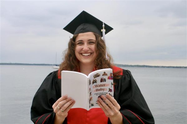 A smiling young woman in a graduation cap and gown holds a paperback book