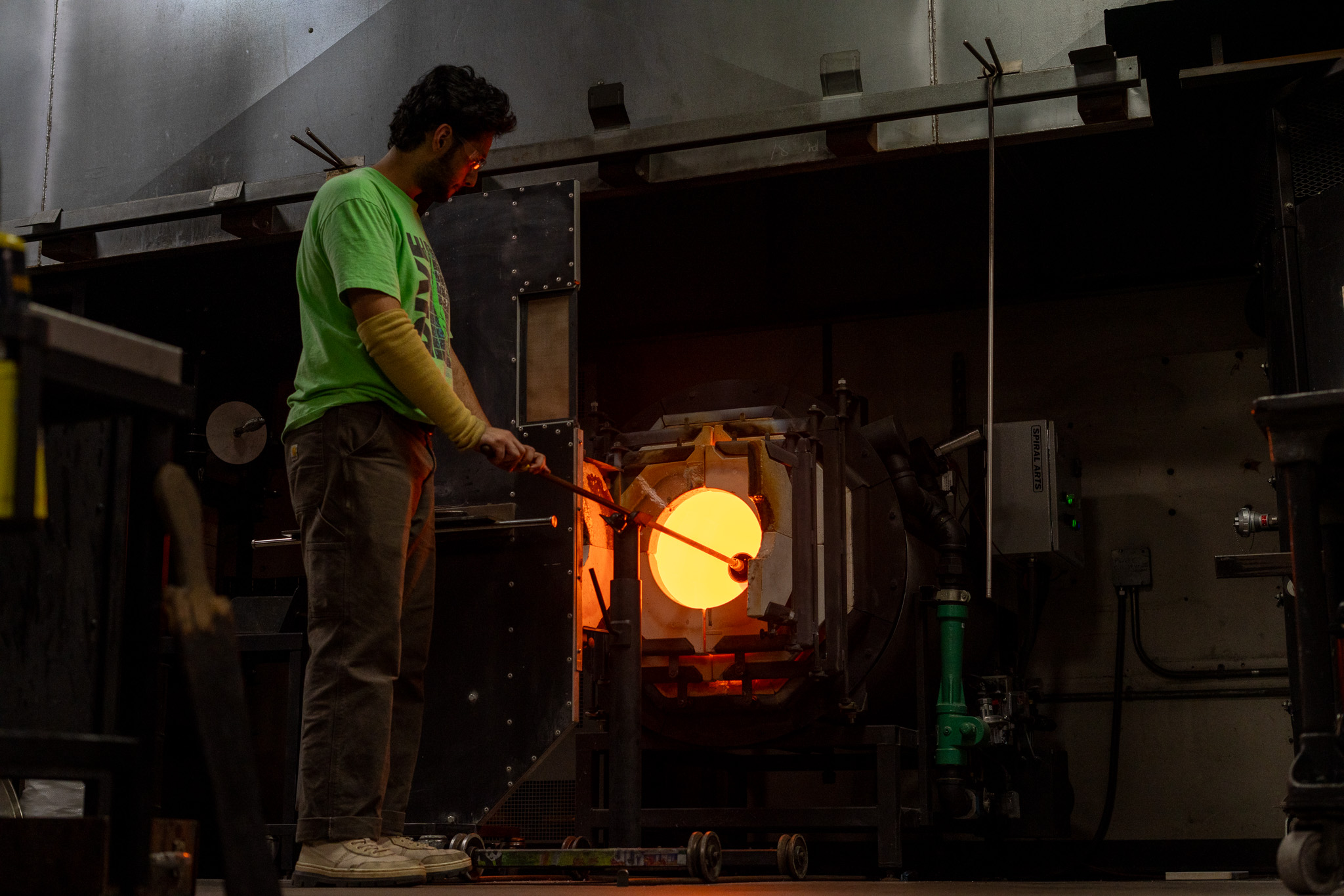 a man in a glass blowing lab stands in the orange glow of an open furnace