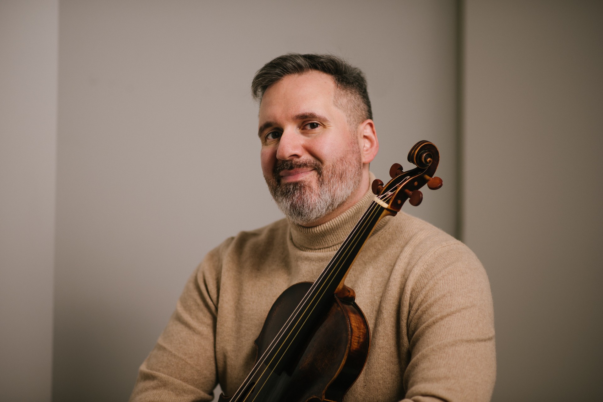 A man in a camel colored sweater is holding a viola while looking directly at the camera.