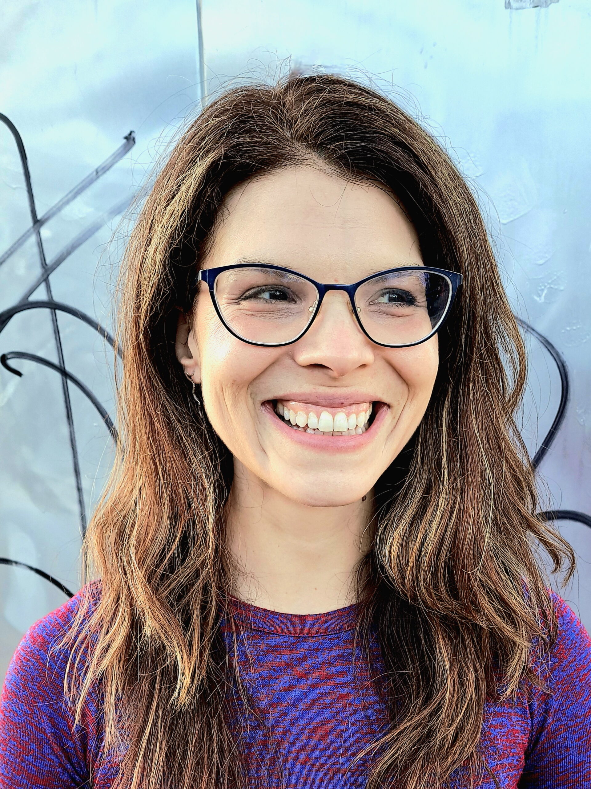 A woman with long brown hair wearing a purple and red sweater and glasses smiles at something off camera