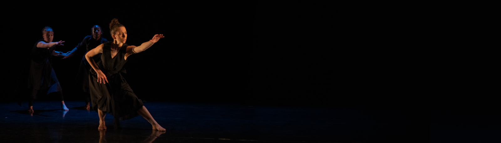 three dancers wearing black dresses on a dark stage, reaching dramatically with their arms