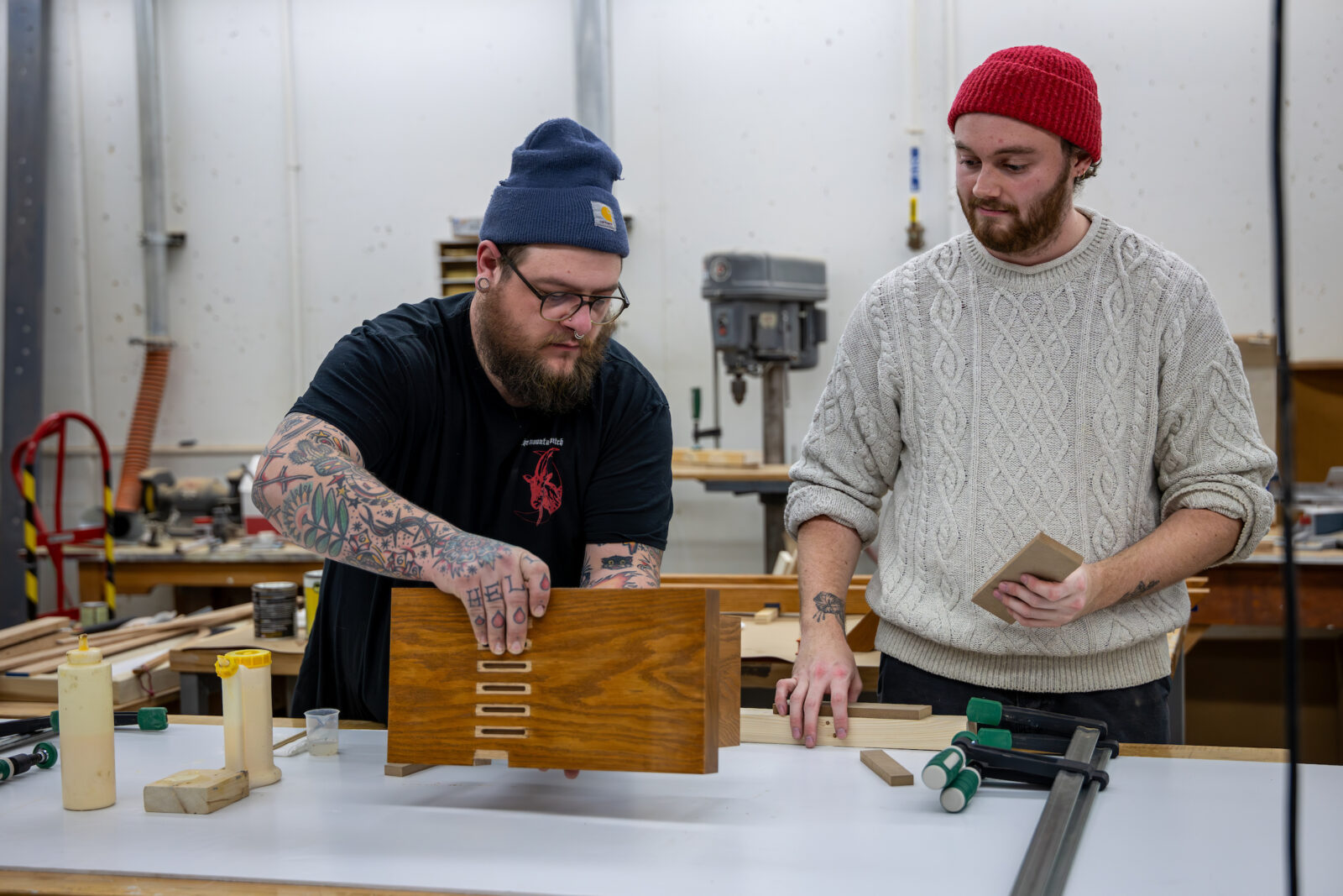 Two men in a workshop holding pieces of wood, with glue and clamps sitting on the workbench