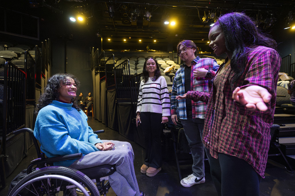 a young woman sitting in a wheelchair smiles at the three college students standing around her speaking animatedly