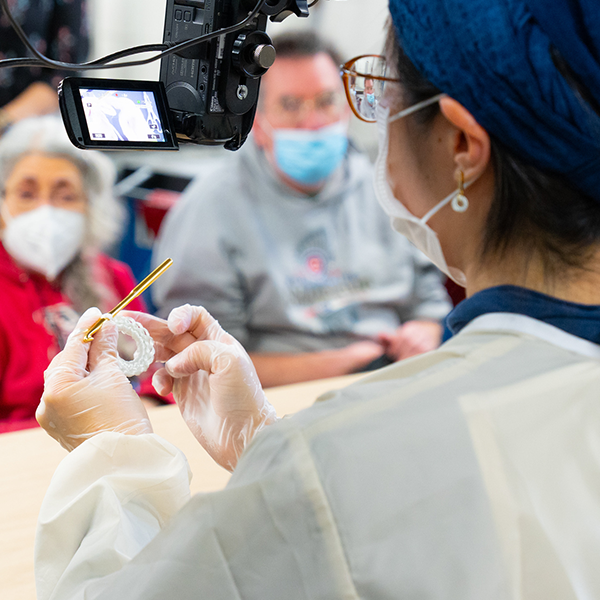an artist in residence in surgical gown and gloves, crochets a delicate white loop in front of an audience while being filmed