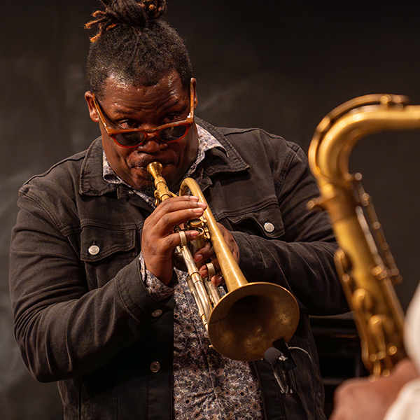 an artist in residence wearing a black denim jacket and sun glasses plays the trumpet, making eye contact with a saxophone player, off camera