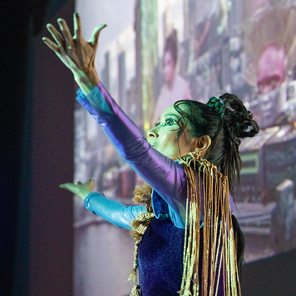 an artist in residence in a futuristic purple outfit with long gold fringe, gestures with arms wide while speaking to an audience