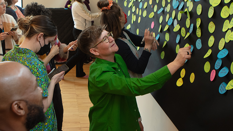 a woman in a green shirt reads colorful sticky notes posted all over a black board on the wall