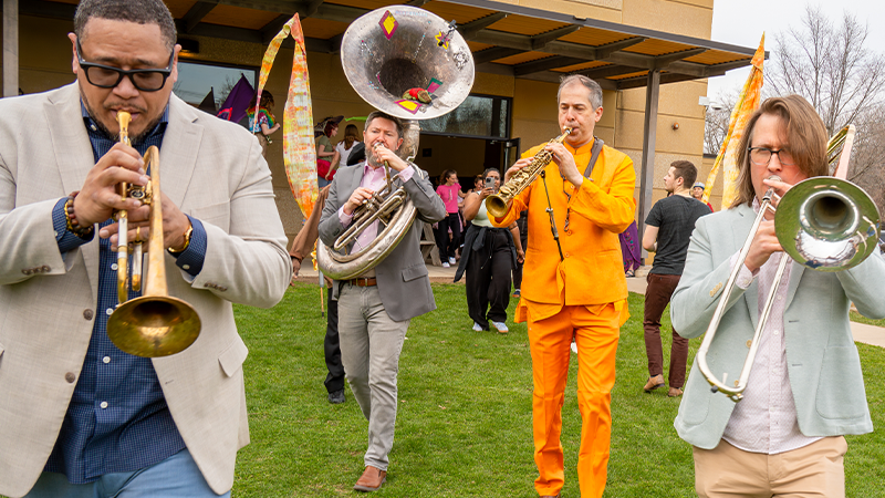 four musicians playing brass instruments as they leave a building and walk out onto grass