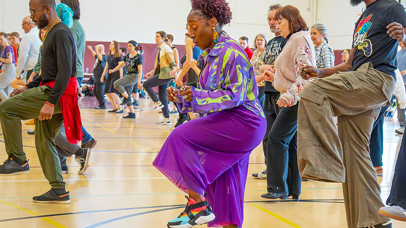 a woman in a vibrant purple outfit leads a gymnasium of dancers