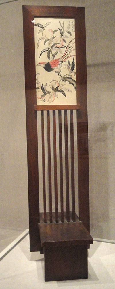 photograph of an original Frank Lloyd Wright frame- a tall slender wooden construction with a Japanese print framed at the top with wooden slats underneath, and a bench-like base to allow the frame to stand unsupported