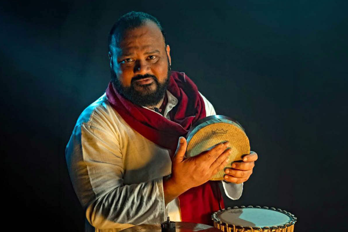 A musician holding a kanjira, a traditional South Indian frame drum, stands with other percussion instruments against a dark background