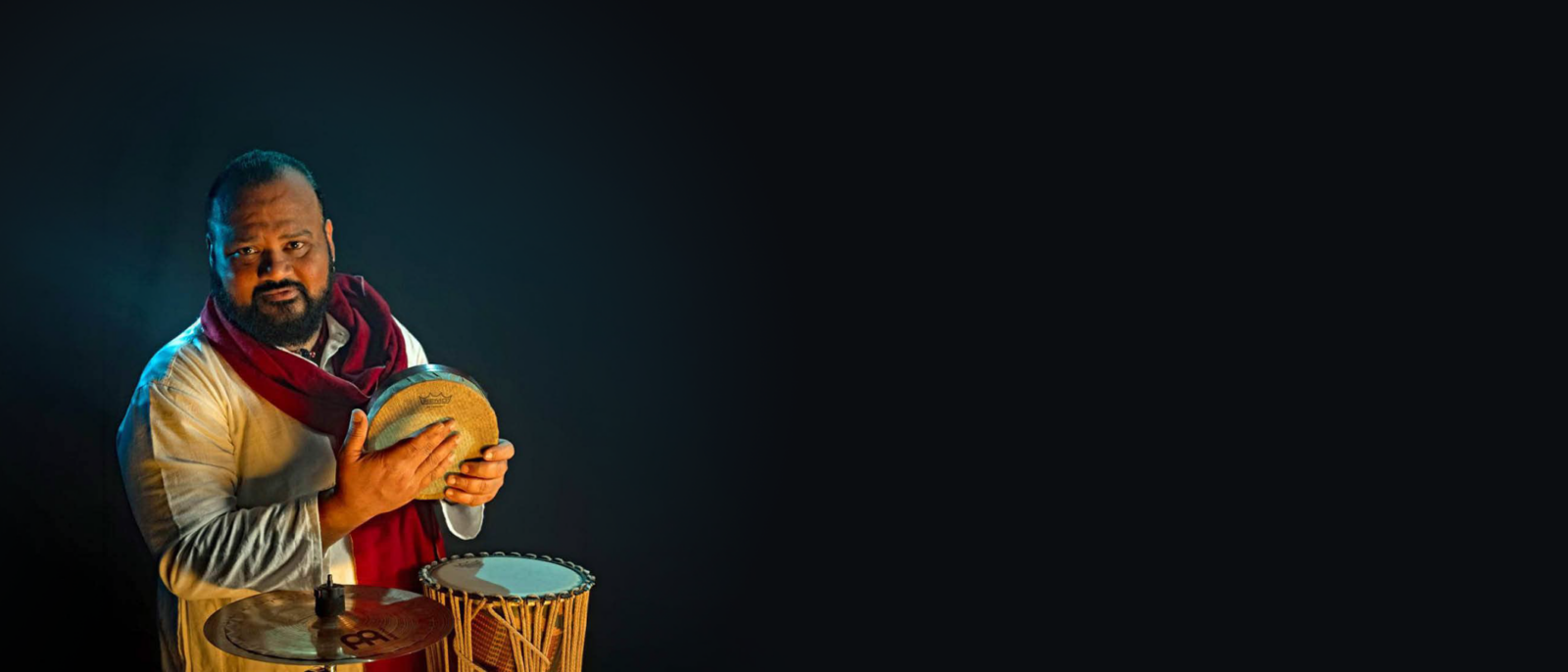 A musician holding a kanjira, a traditional South Indian frame drum, stands with other percussion instruments against a dark background
