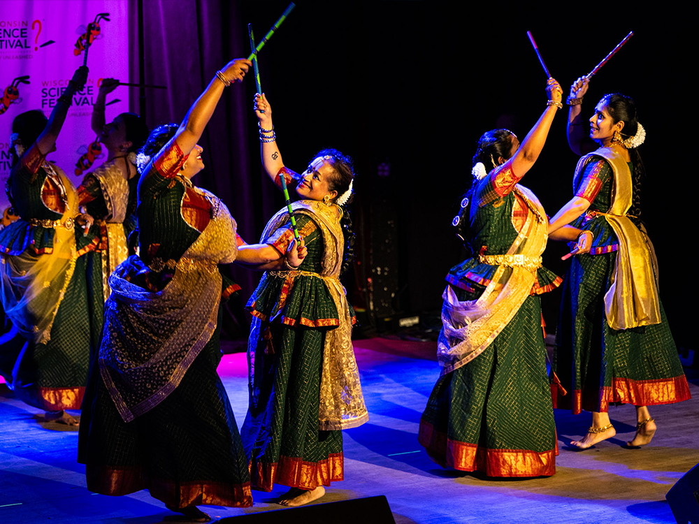 six women in traditional indian dresses dancing on a stage with sticks called dandiyas in both hands