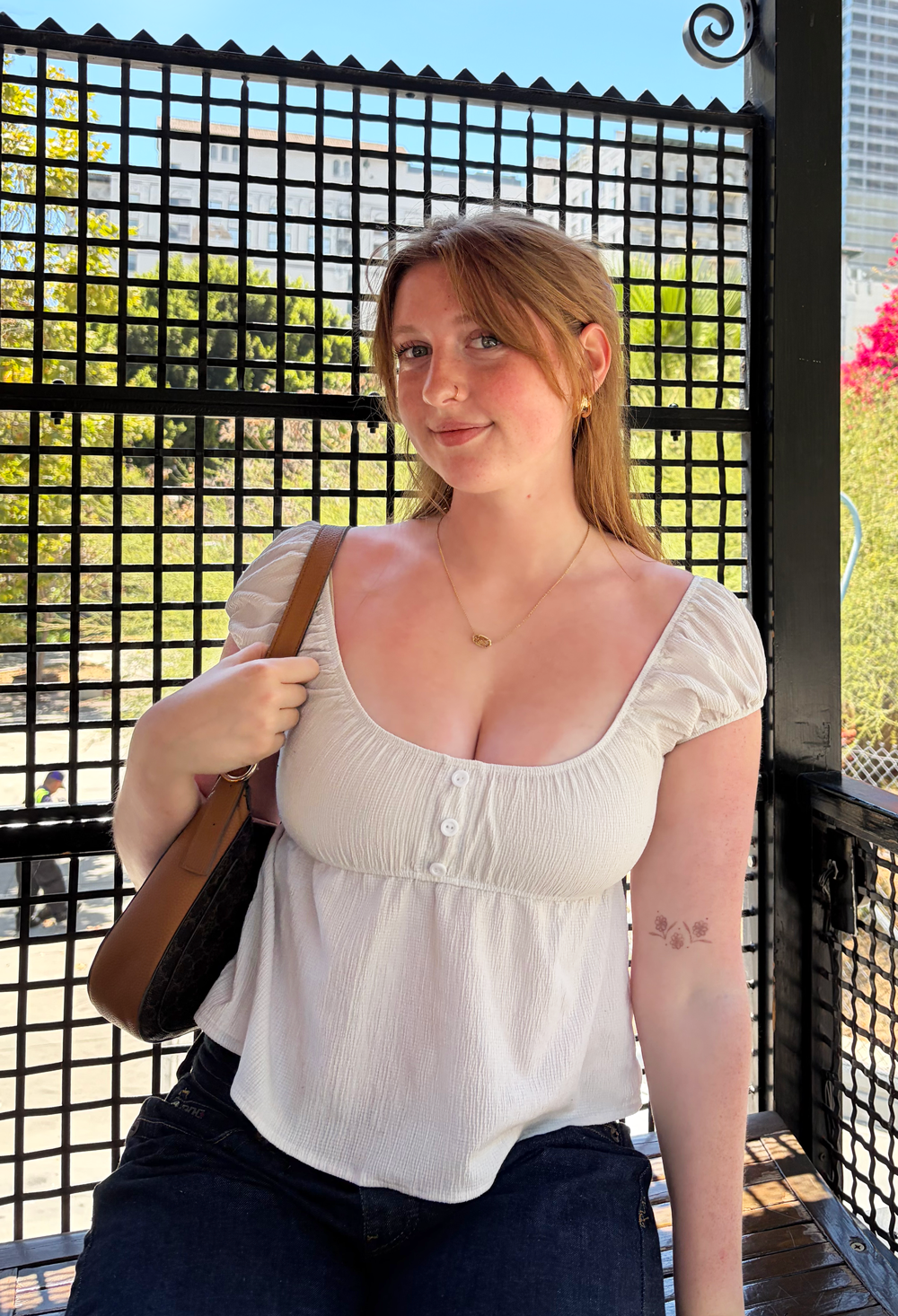 a young woman wearing a white top standing in front of a metal grate, with trees in the background